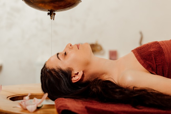 side view of relaxed young woman lying under shirodhara vessel during ayurvedic procedure